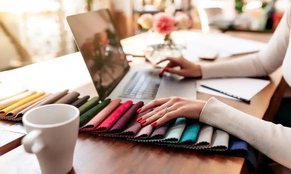A large swatch of upholstery cover choices on a desk with a woman touching the samples while on her laptop